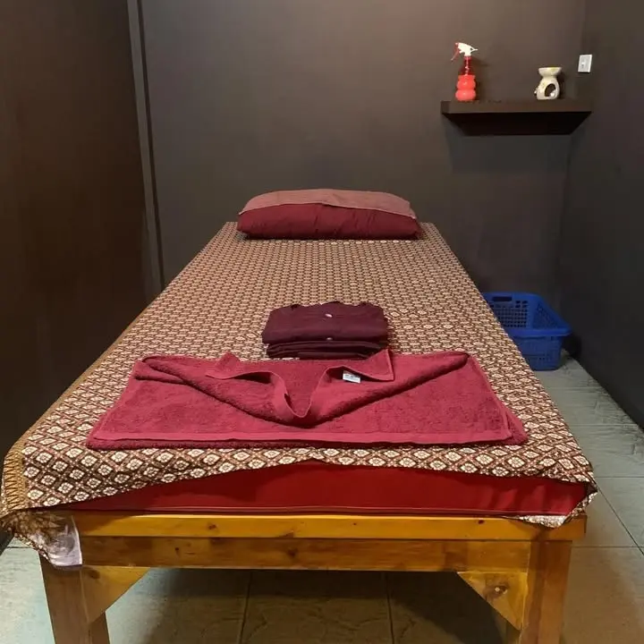 A private massage room: wooden massage bed with Thai-weave cover, burgundy pillow, towel and folded massage clothes